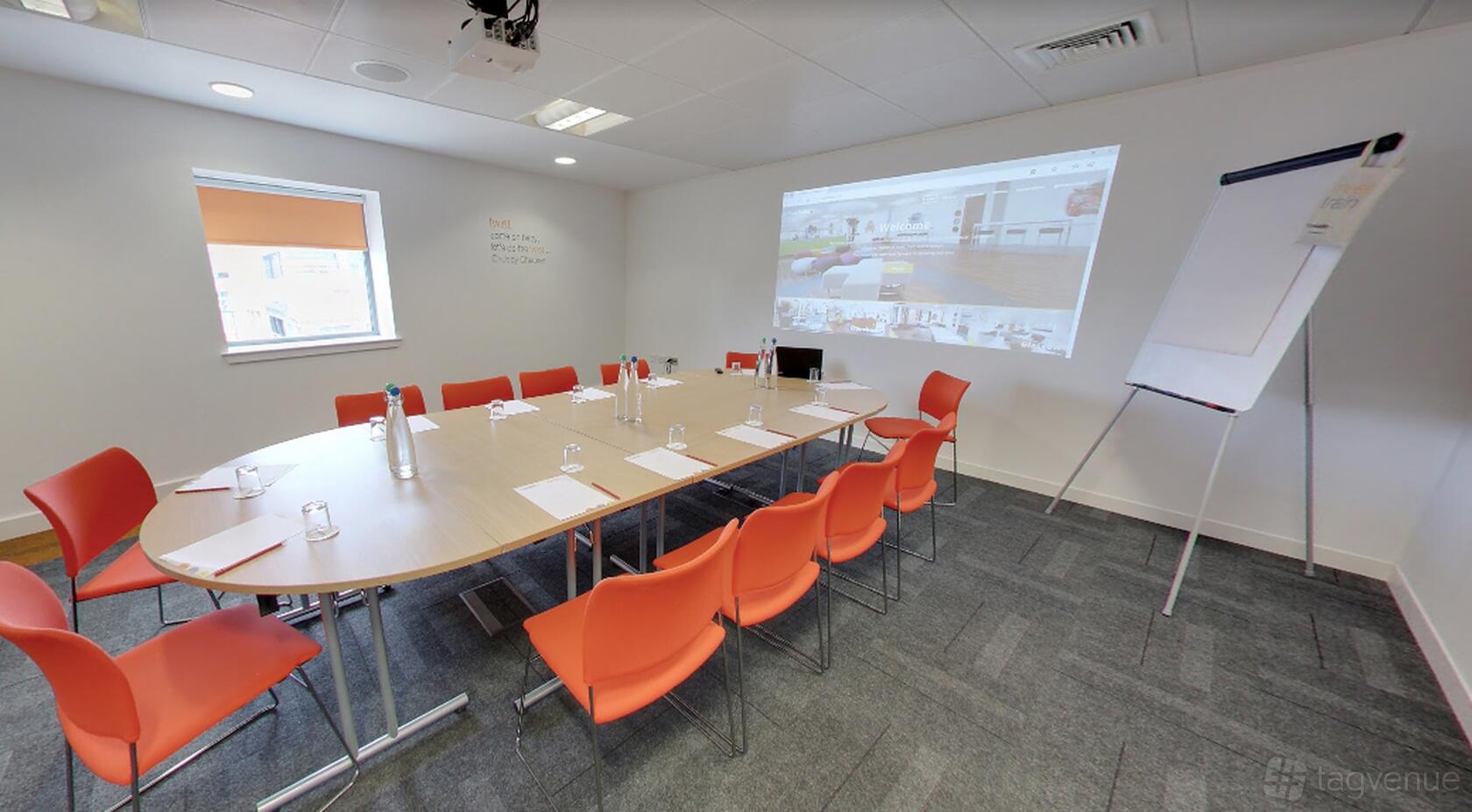 A conference centre with an oval table, orange chairs, projector screen, and flip chart at The Studio Glasgow.