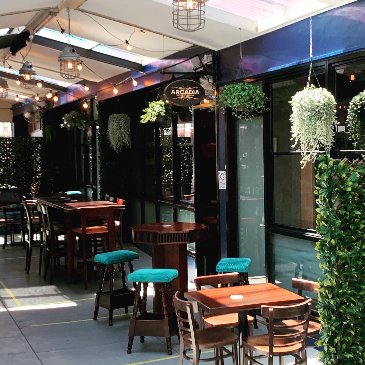 An outdoor bar with wooden tables, teal barstools, and hanging greenery at Arcadia Whisky Lounge.