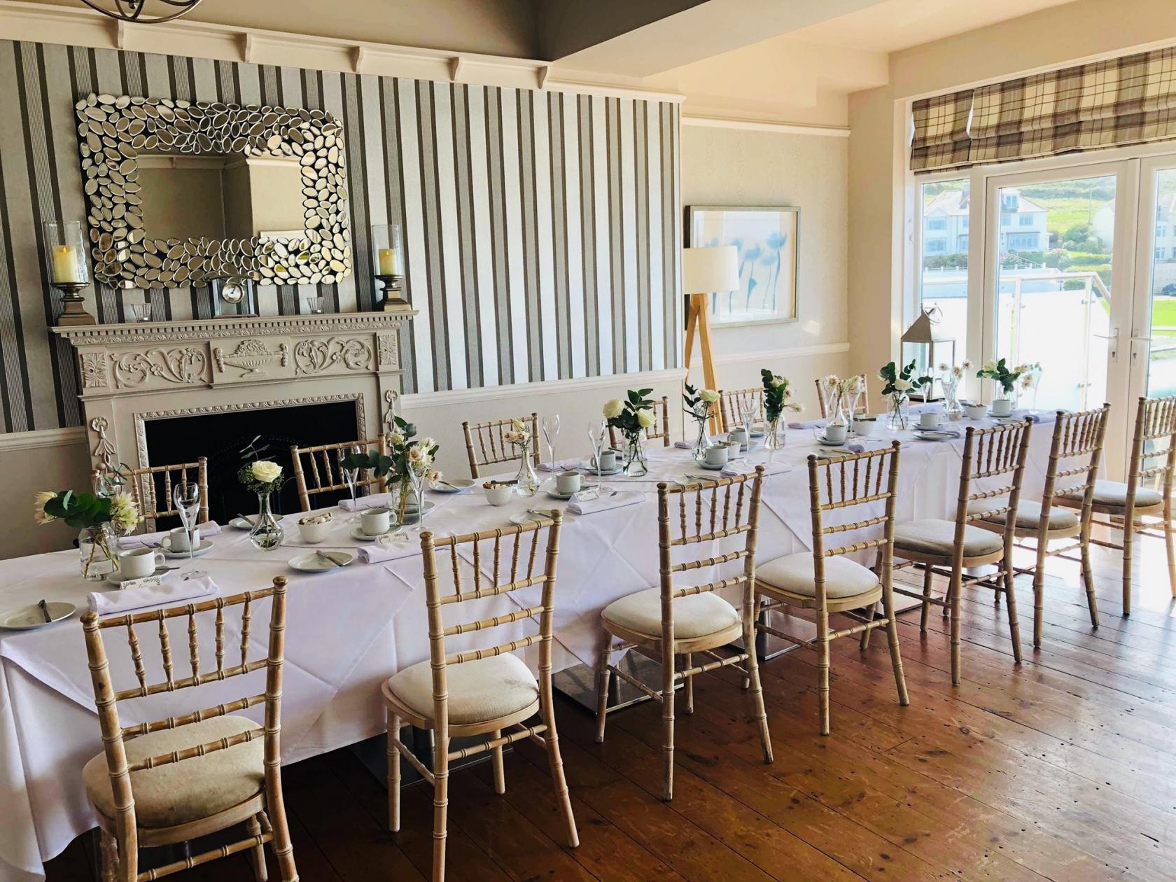 An event space with a long rectangular table set with white linens and wooden chairs near a decorative fireplace at Watersmeet Hotel.