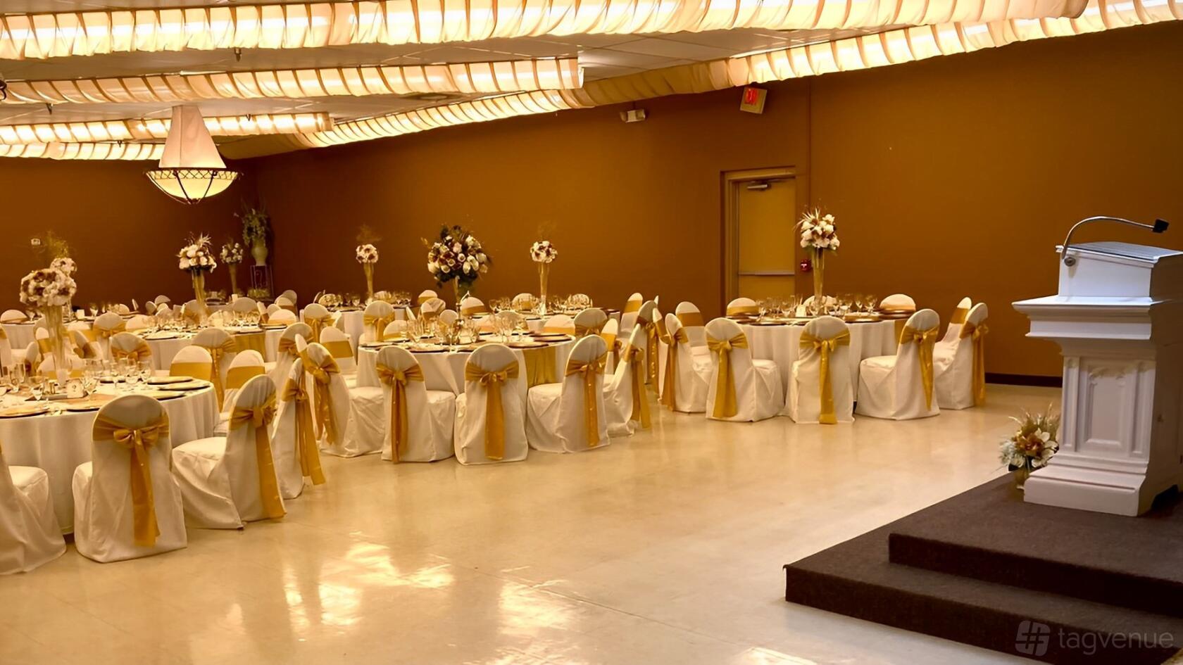 A ballroom with round tables draped in white linens and gold chair sashes, floral centerpieces, and a podium at JS Venue Plus.