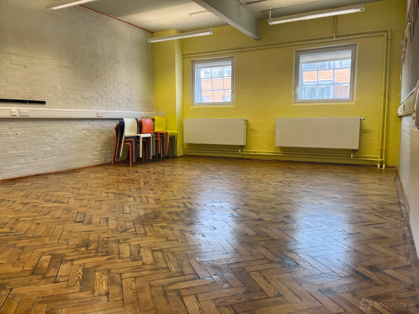 A rehearsal room with parquet wood flooring, bright yellow walls, and stacked colorful chairs at Collective Acting Studio.