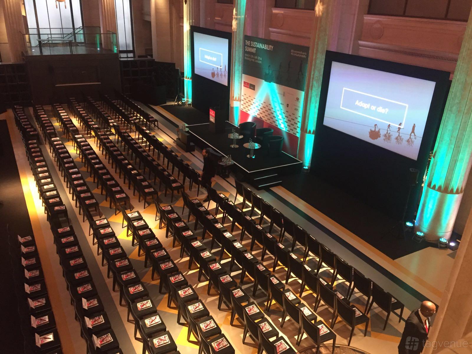 A grand hall with rows of black chairs, two large projector screens, and tall columns with uplighting at Banking Hall.