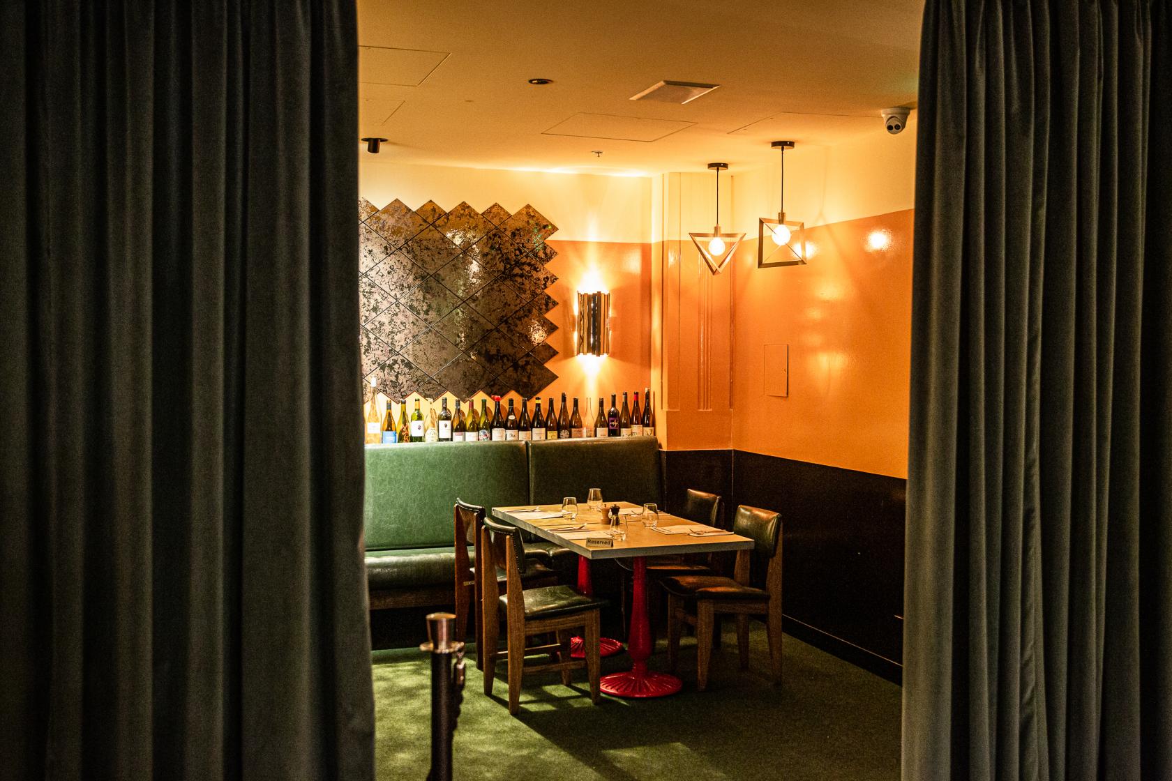 A dining room with green velvet banquette seating, patterned wall art, and bottles displayed on a shelf at Natural History Public Bar.
