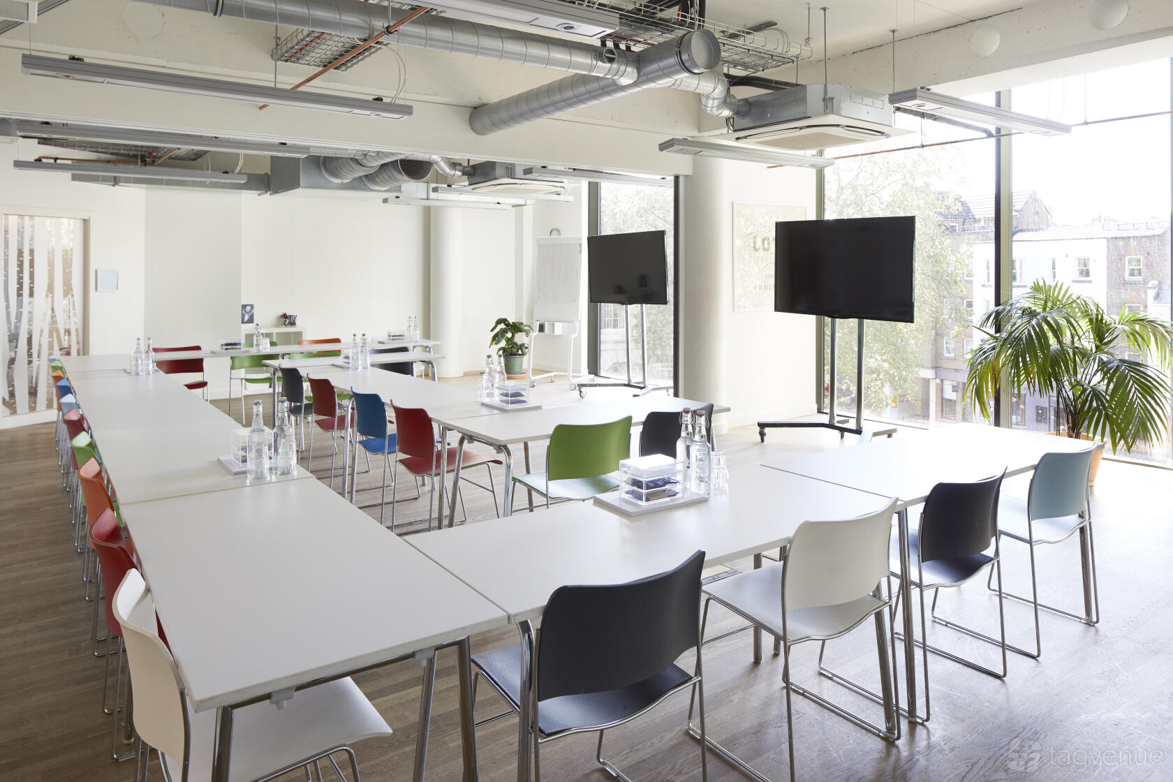 An auditorium with large windows, colorful chairs, long white tables, and exposed ductwork at Wallacespace Clerkenwell Green.