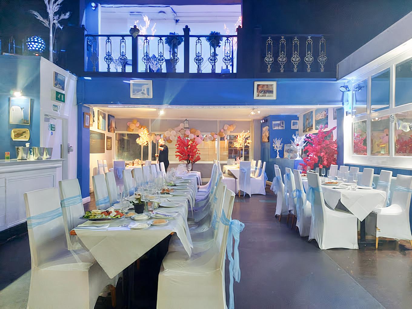 An event space in a restaurant with white chairs, long banquet tables, blue walls, and decorative red centerpieces at Santorini Greek Restaurant.
