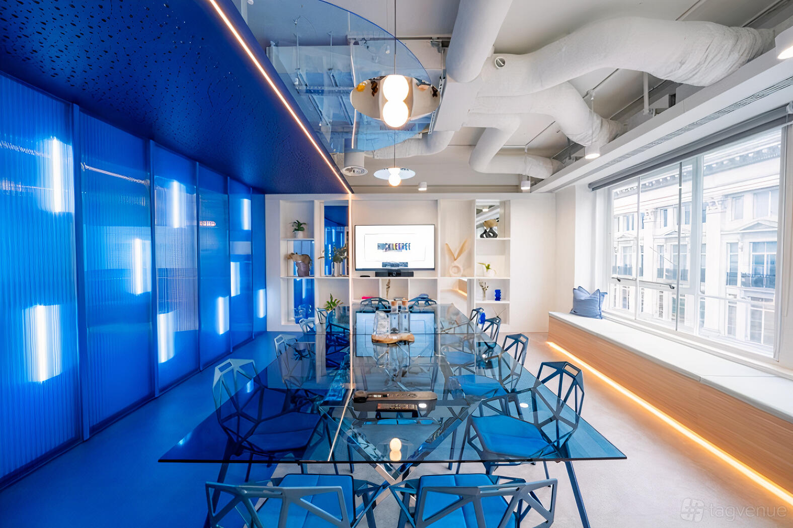 A meeting room with a glass table, geometric blue chairs, floor-to-ceiling windows, and accent lighting at Huckletree Oxford Circus.