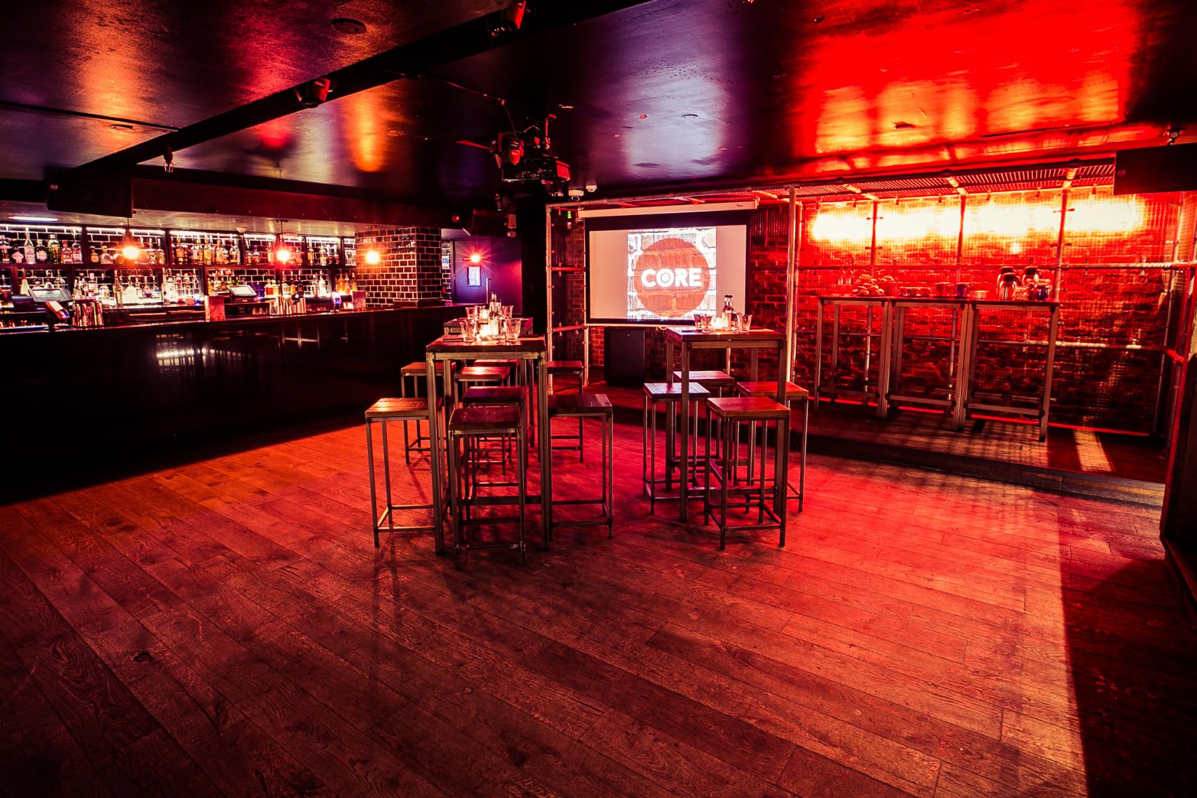 An event space in a bar with exposed brick walls, a large projection screen, and high tables at Core.