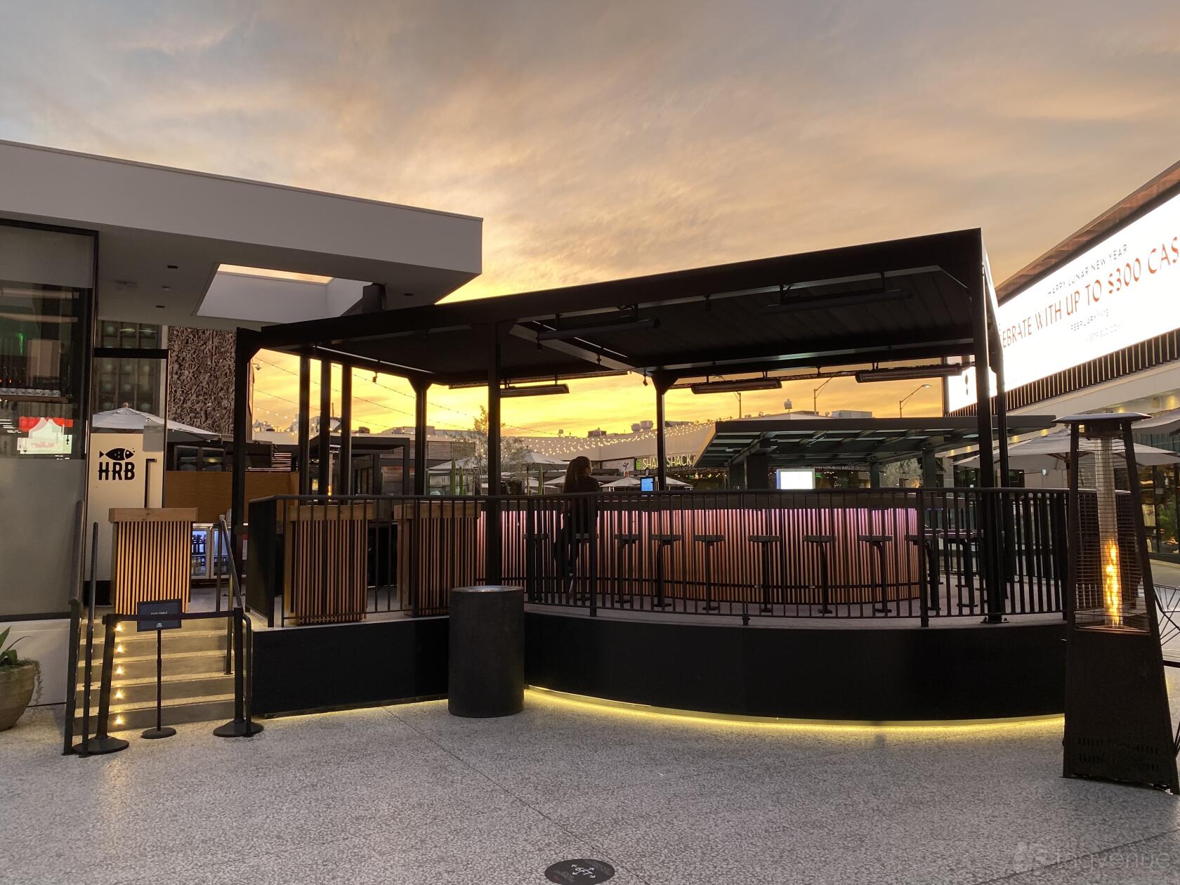 An outdoor restaurant area with a covered patio, perimeter railing, and warm sunset lighting at HRB Experience.