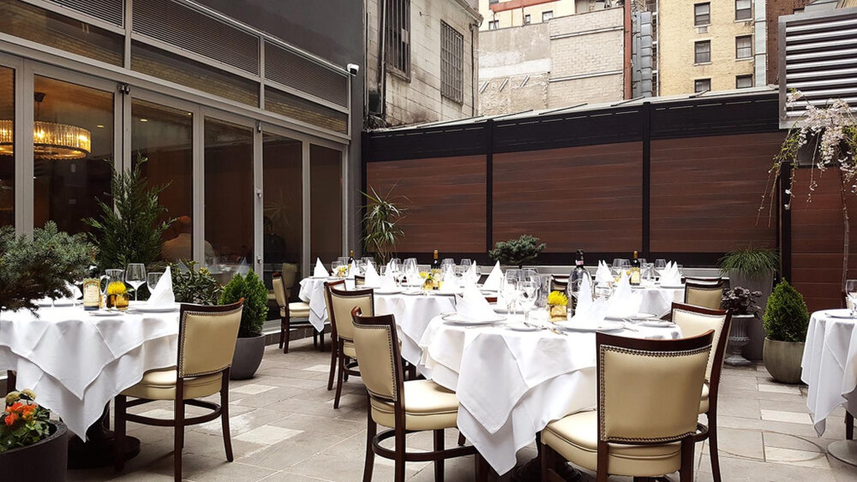 An outdoor dining area with round tables draped in white linens and potted plants at Allora.