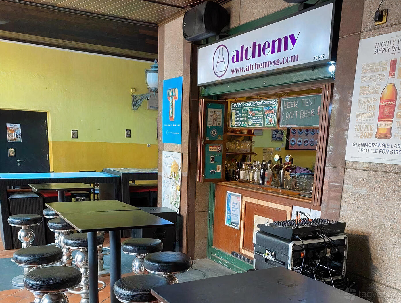 A pub with high-top tables, round metal stools, and a bar counter under an Alchemy sign