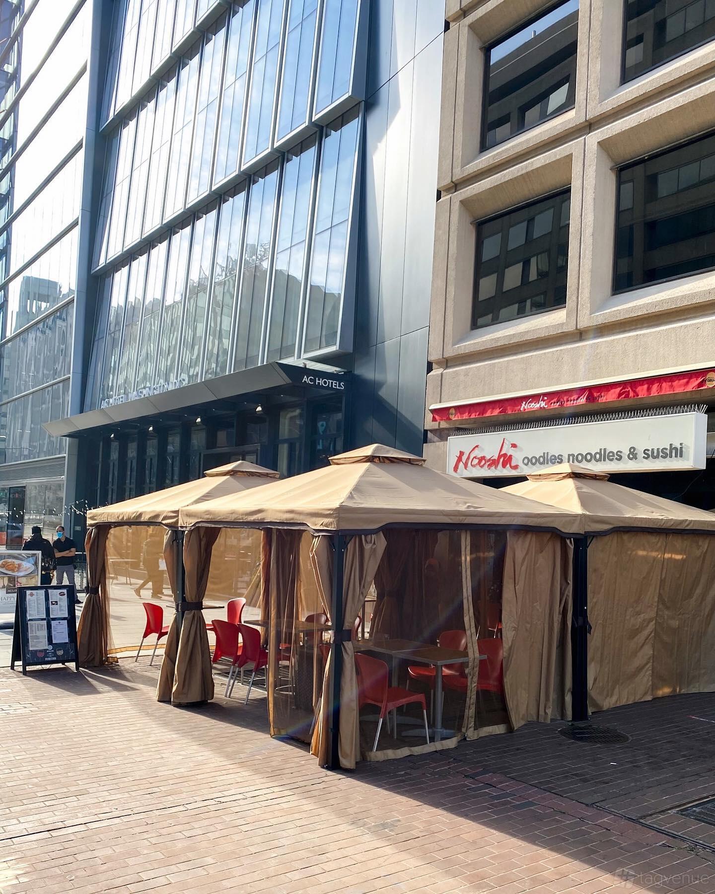 A restaurant with outdoor seating under tan canopies and red chairs in front of Nooshi 19th Street.
