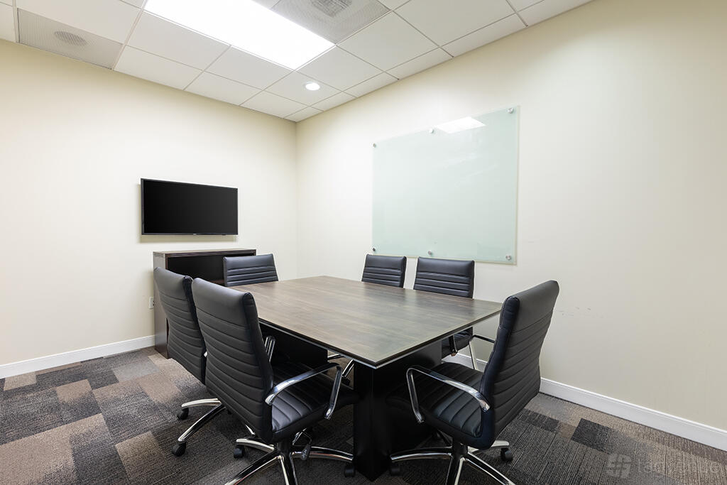 A coworking space meeting room with a rectangular table, six leather chairs, wall-mounted TV, and a glass whiteboard at Premier Workspaces - Carlsbad.