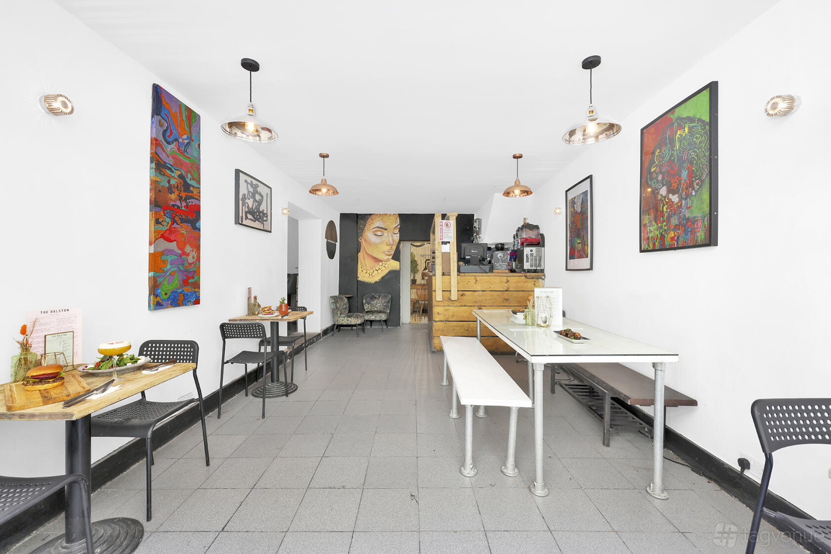 A bar with colorful wall art, pendant lighting, and a mix of wooden benches and metal chairs at The Dalston Lounge.