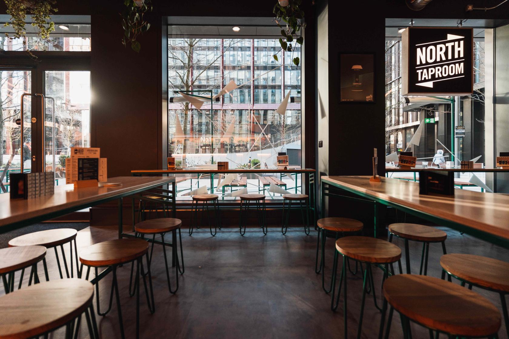 A bar with large windows, round wooden stools, and high tables at North Bar Circle Square.
