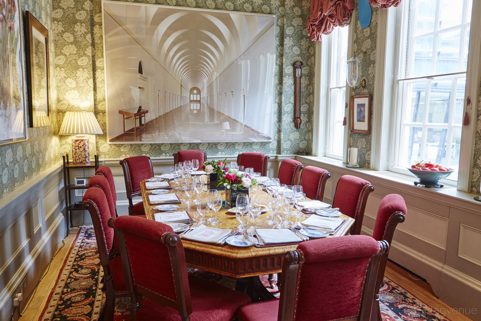 A dining room with red upholstered chairs, wood table, patterned wallpaper, and large windows at The Walbrook Club