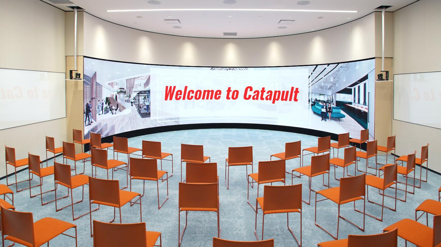 An auditorium with tiered orange chairs and a large curved digital screen at Catapult.