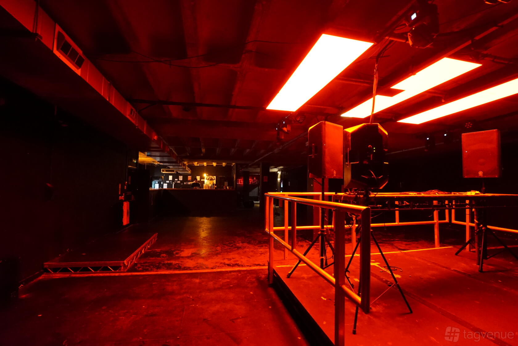 An event venue with red ceiling lights, exposed concrete beams, and a metal-railed stage area at Clock Factory.