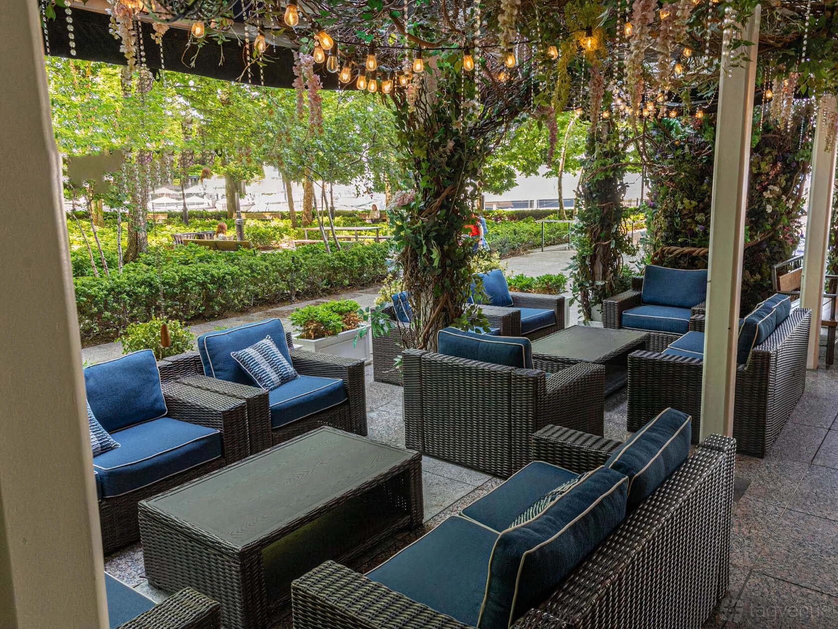 A lounge with wicker seating, blue cushions, and hanging string lights overlooking a garden at Le District.