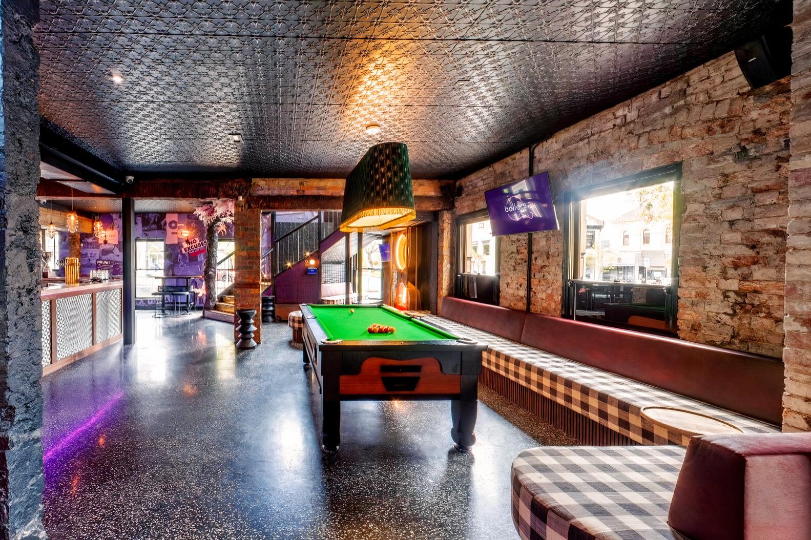 An event space in a bar with a central pool table, exposed brick walls, patterned banquette seating, and large windows at Ballers Clubhouse Carlton.