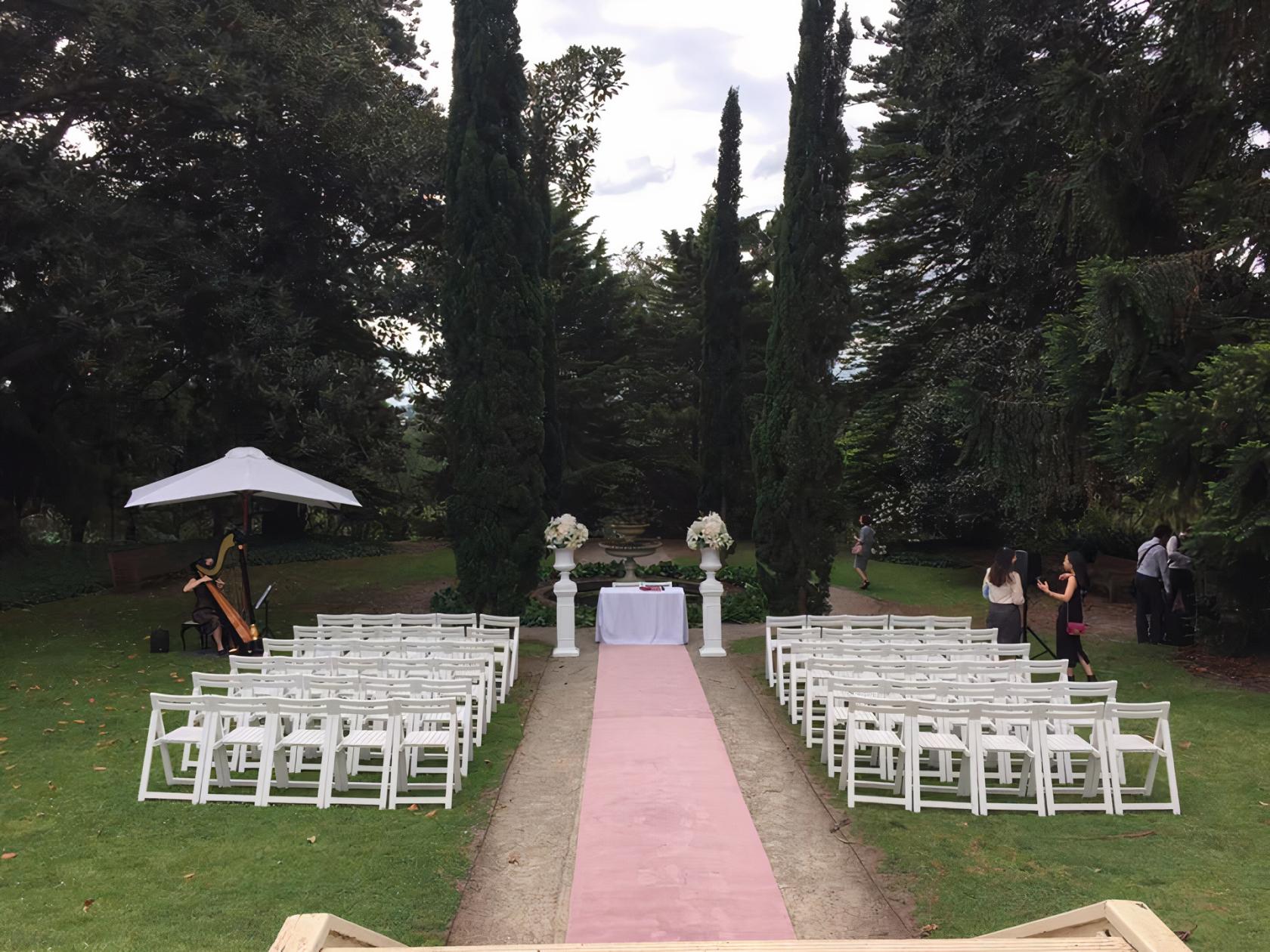 An outdoor ceremony area with white chairs, floral arrangements, and tall trees at Como House and Garden.