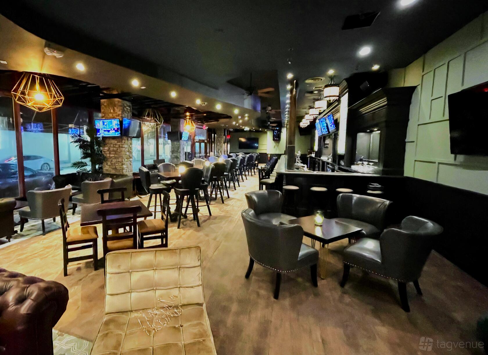 A bar with leather armchairs, high-top tables, large windows, and multiple wall-mounted TVs at Mickey Burkes.