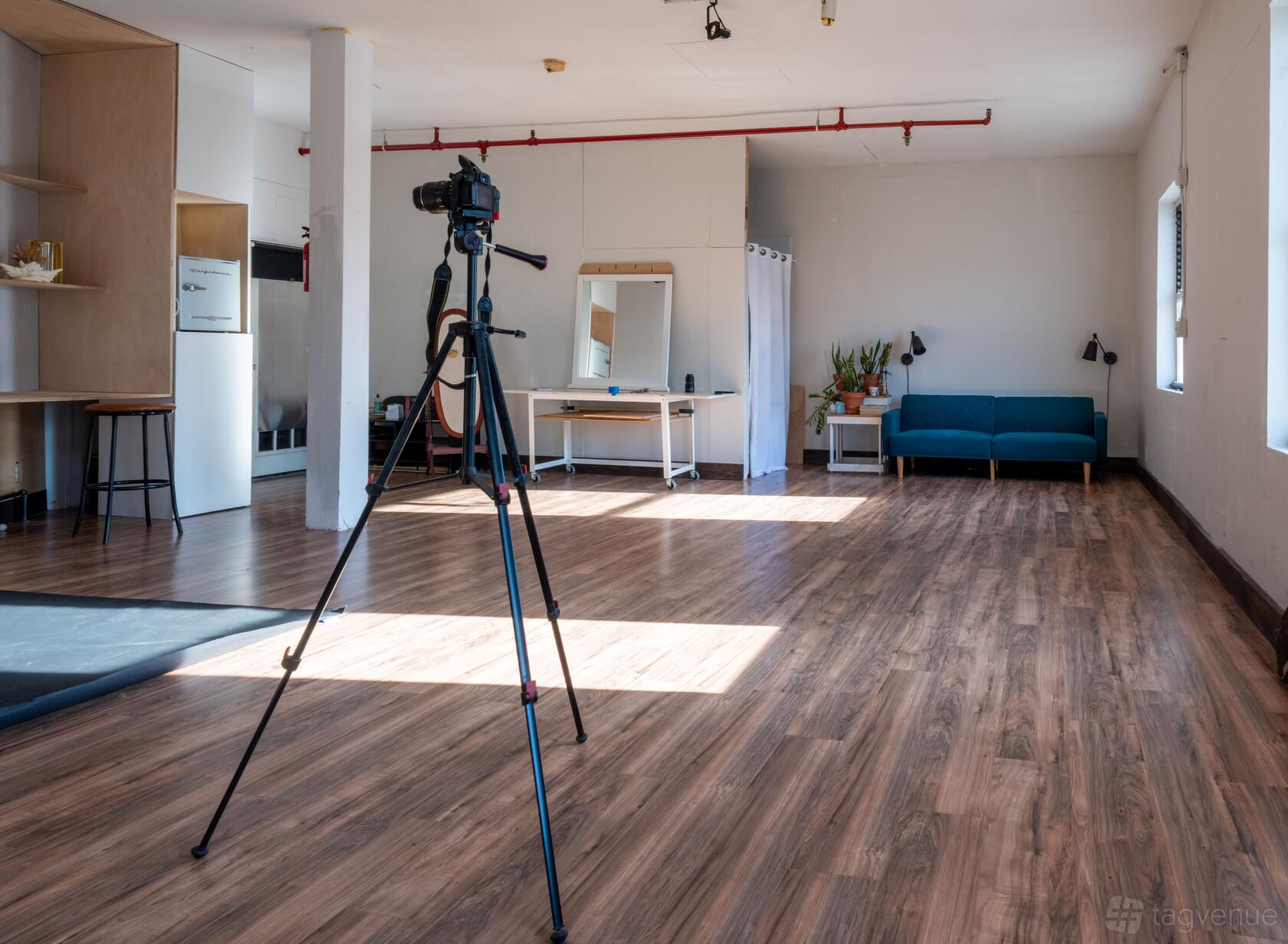 A photo studio with wood floors, a camera on a tripod, a blue sofa, and a makeup station at The Greenpoint Gallery.