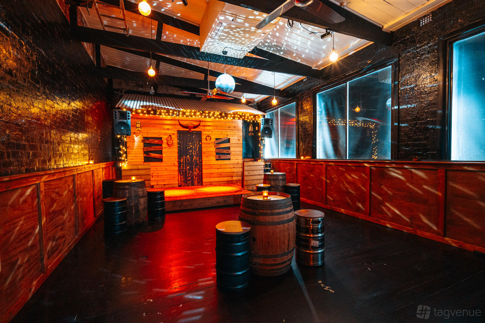 A function room in a bar with a small wooden stage, barrel tables, and string lights at Death or Glory.
