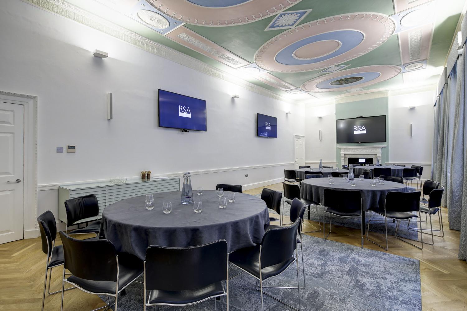 A meeting room with round tables, black chairs, ornate pastel ceiling, and wall-mounted screens at RSA House.