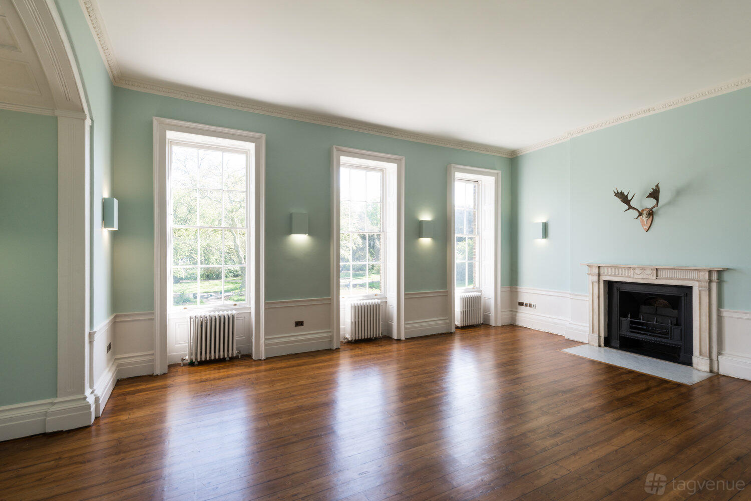 A dining room with large sash windows, hardwood floors, pale blue walls, and a decorative fireplace at Clissold House.