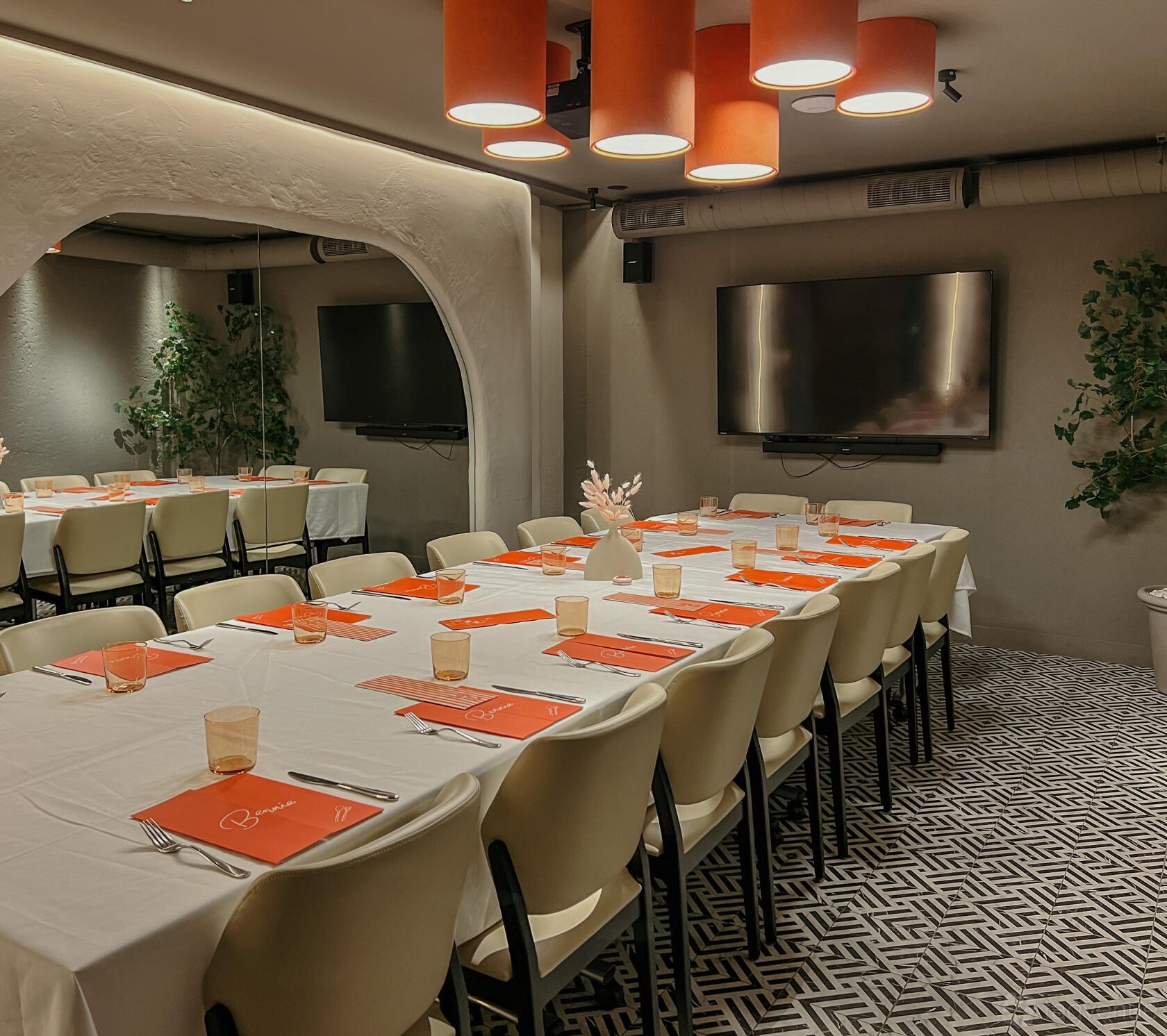 A private dining room with long tables covered in white linens, orange placemats, and a wall-mounted TV at Bennie Restaurant.