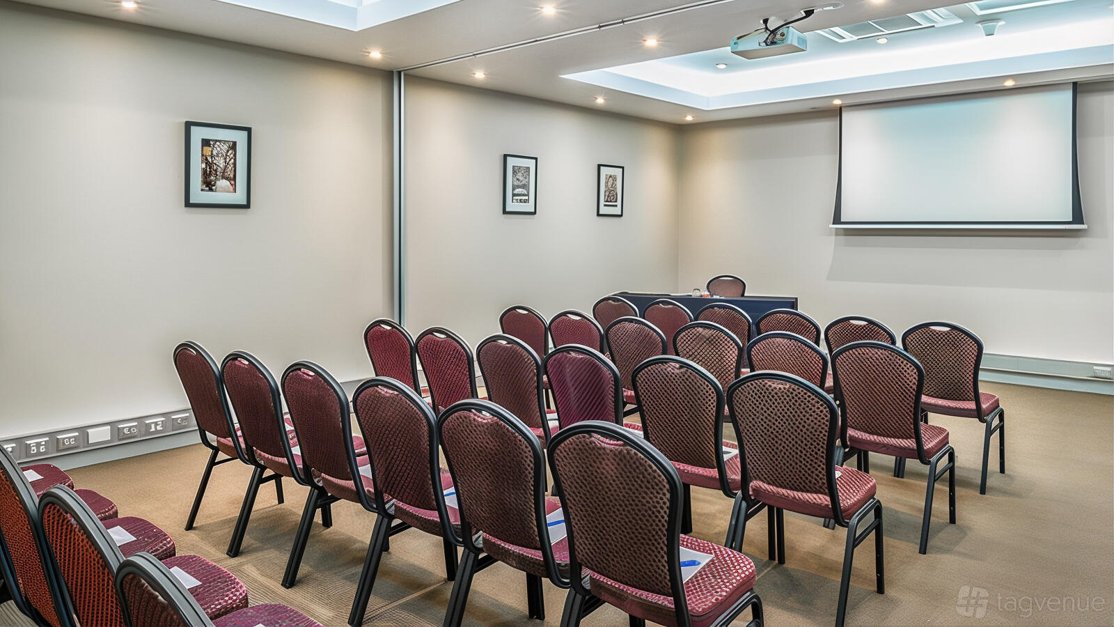 A function room with rows of red cushioned chairs, wall art, and a ceiling projector at Mantra Melbourne Airport.