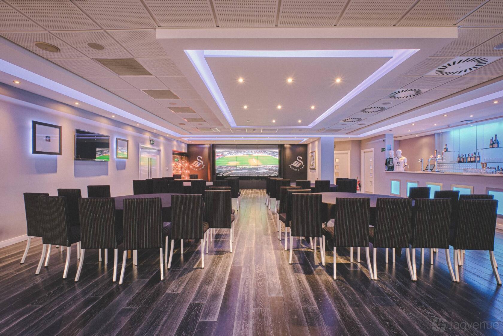 A function room with rows of black chairs, wood flooring, and ceiling lights facing a large screen at Swansea.com Stadium.
