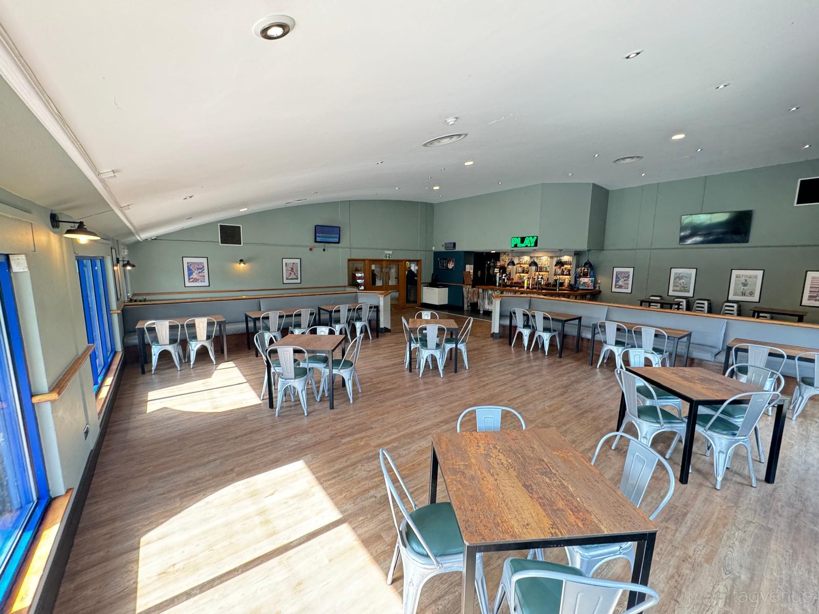 A function room with metal chairs, wooden tables, a long bar, and large windows at Power League Newham.