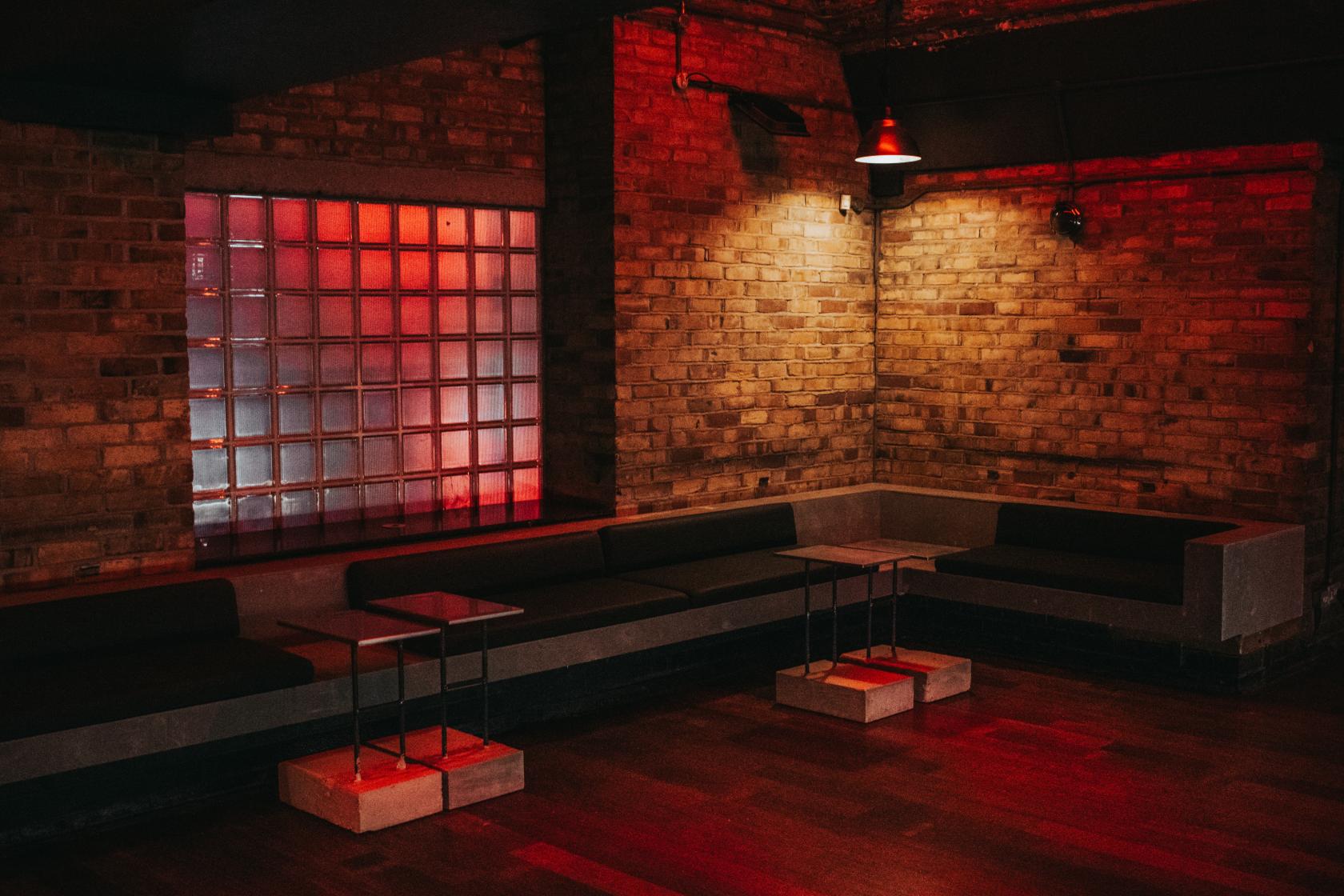 A lounge with brick walls, built-in bench seating, and small square tables under red lighting at fabric industrial arts events venue.