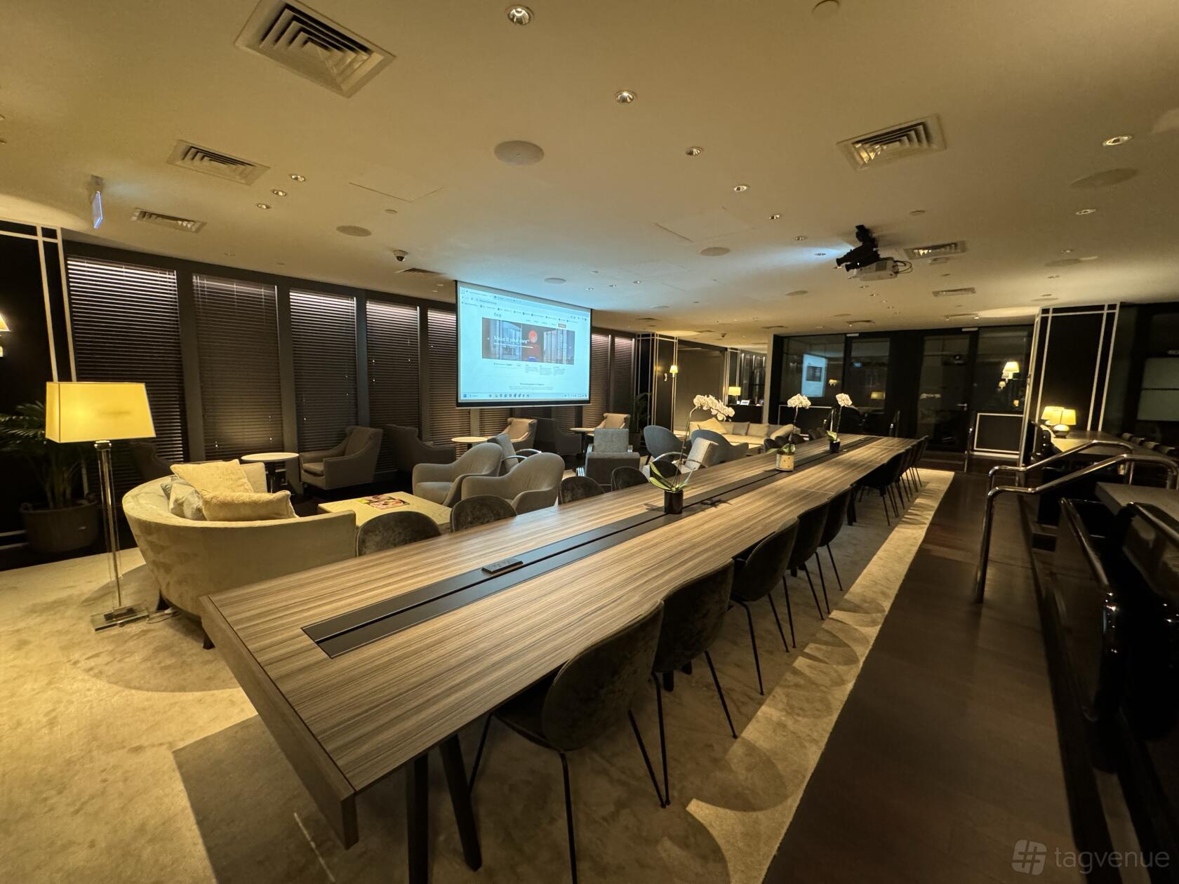 An event space with a long communal table, cushioned armchairs, floor lamps, and a projector screen at The Work Project OUE Downtown.