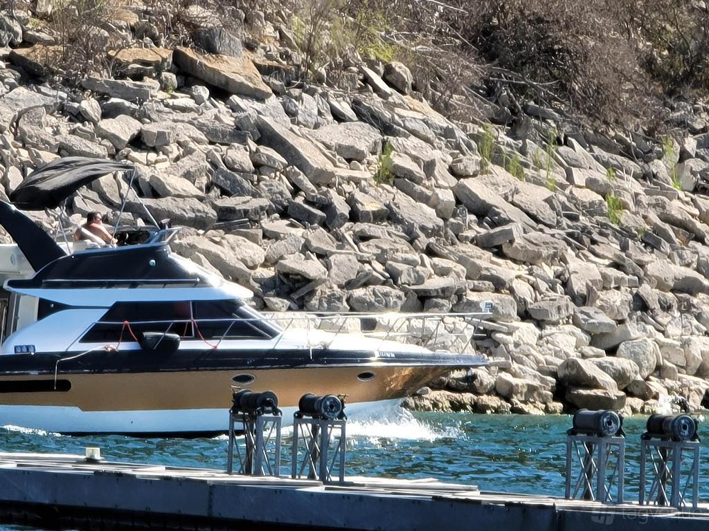 A yacht with a shaded upper deck cruising near a rocky shoreline at Lake Travis Yacht Charters.
