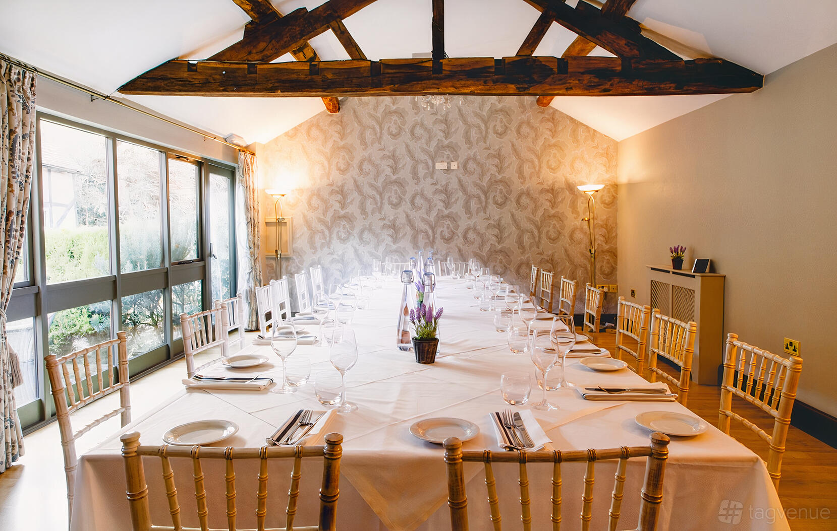 An event venue room with exposed wooden beams, patterned wallpaper, and a U-shaped set of tables at Old Rectory House.