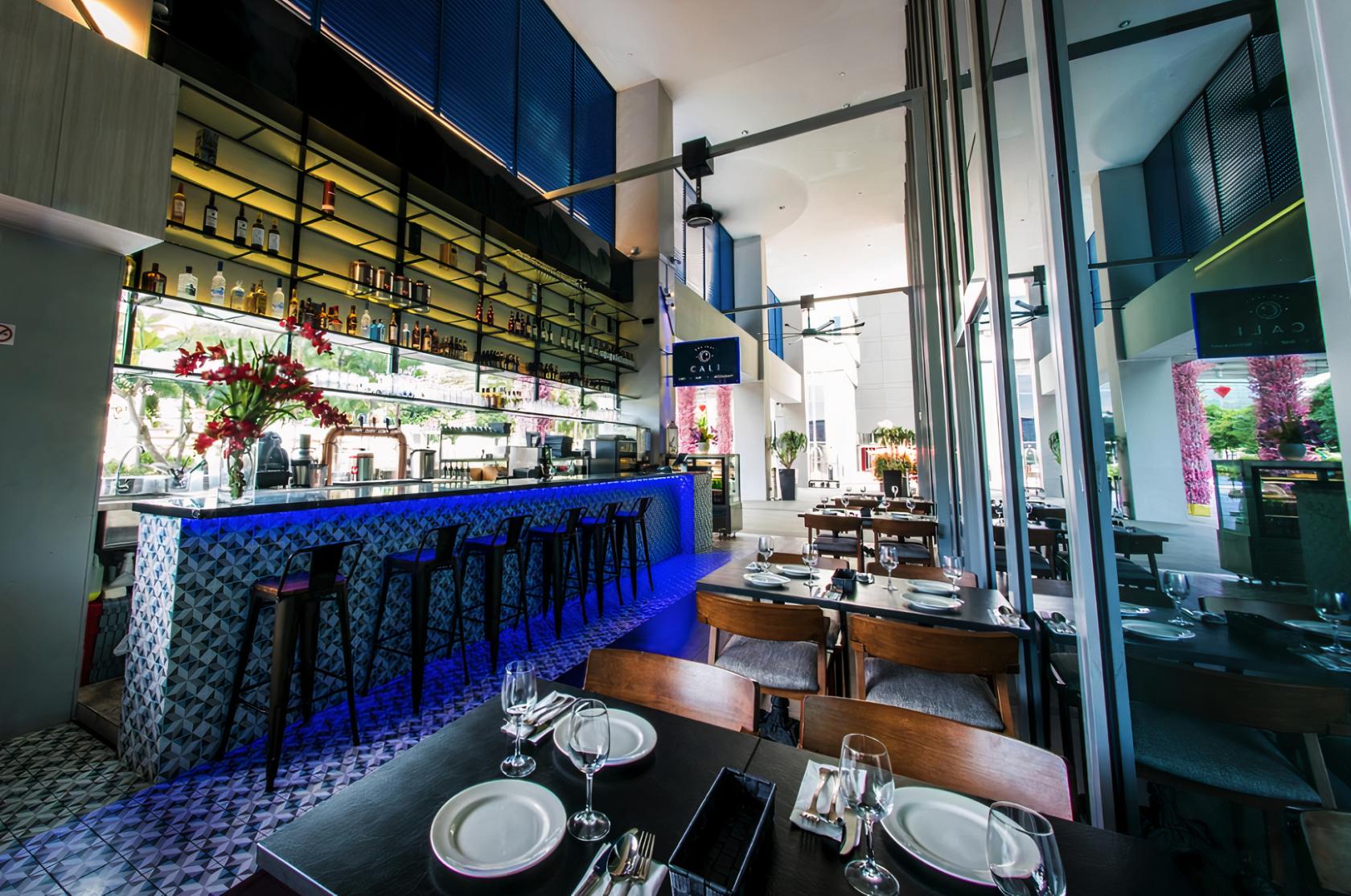 A restaurant with a backlit bar, high ceiling, glass walls, and wooden tables at CALI, Park Avenue Changi.