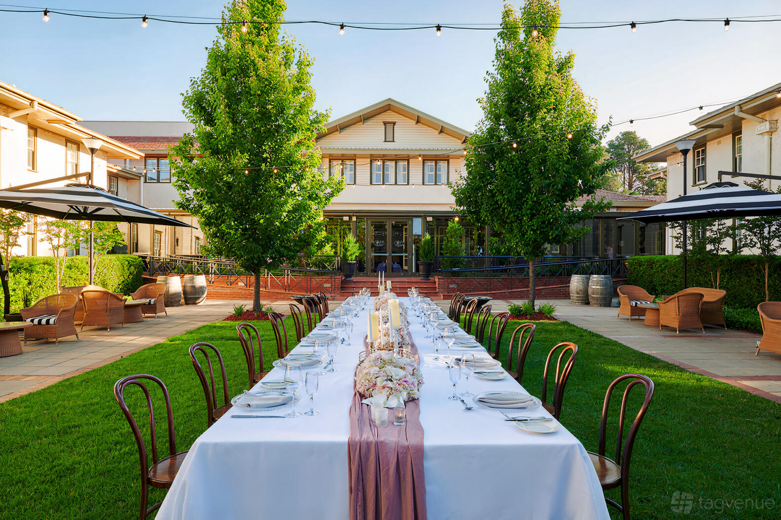 An outdoor hotel courtyard with a long banquet table set on green lawn, string lights, and surrounding trees at Hotel Kurrajong Canberra.