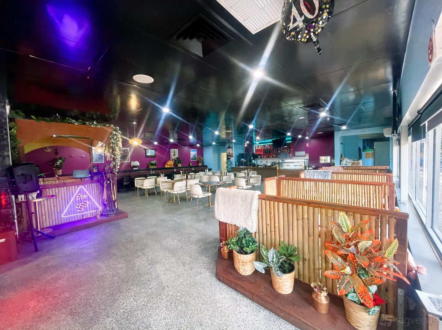 A restaurant interior with bamboo partitions, potted plants, white chairs, neon signs, and bright overhead lighting at Tinkune Momo and Sekuwa House.