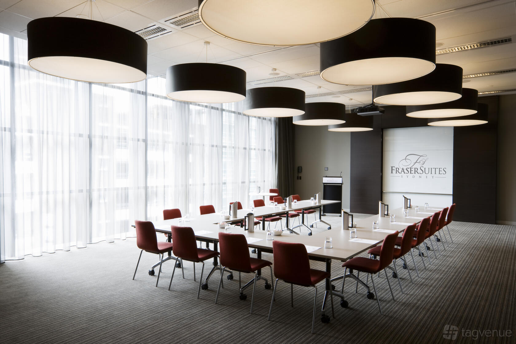 A hotel meeting room with large circular ceiling lights, floor-to-ceiling windows, and U-shaped tables at Fraser Suites Sydney.