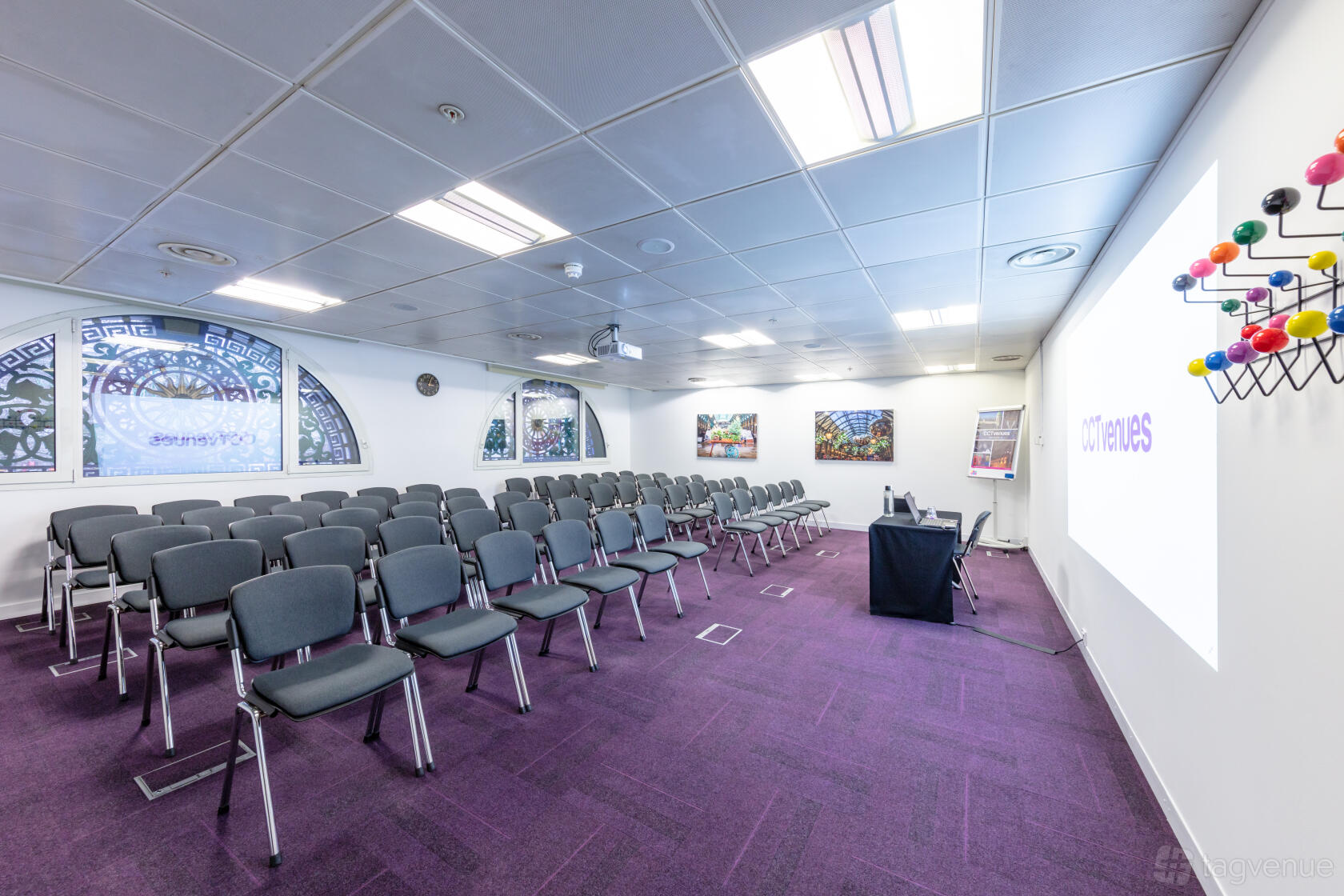 A conference centre with rows of grey chairs, large projector screen, and purple carpet at CCT Venues - Smithfield.