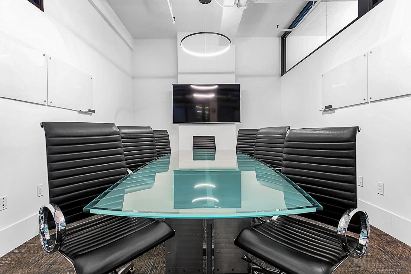 A meeting room with a glass conference table, black leather chairs, wall-mounted TV, and whiteboards at Gravel Road Business Executive Suites.