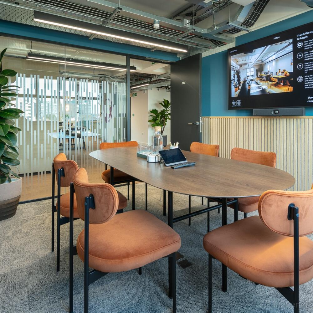 A meeting room with a wooden oval table, orange chairs, glass partition, and wall-mounted screen at Spacemade - Beton.