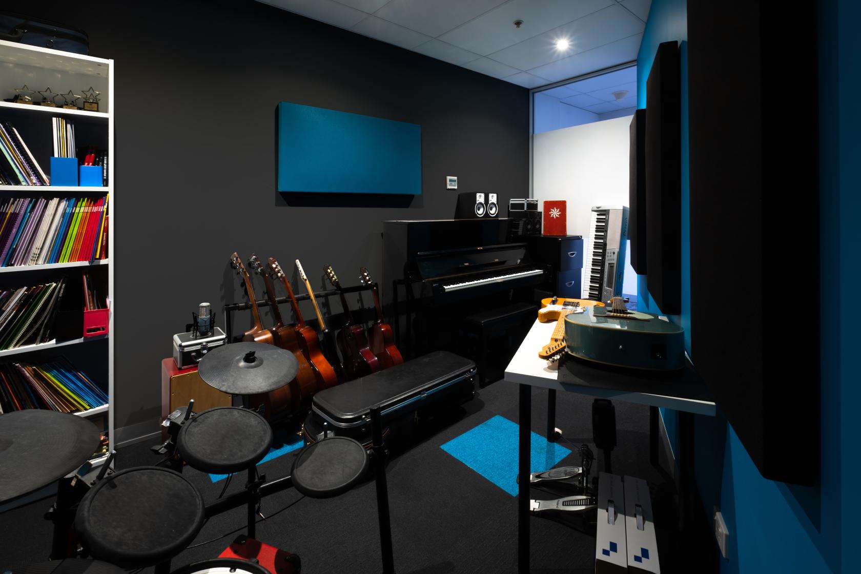 A rehearsal room with a digital drum kit, upright piano, guitars, and soundproofing panels at Music Valley.