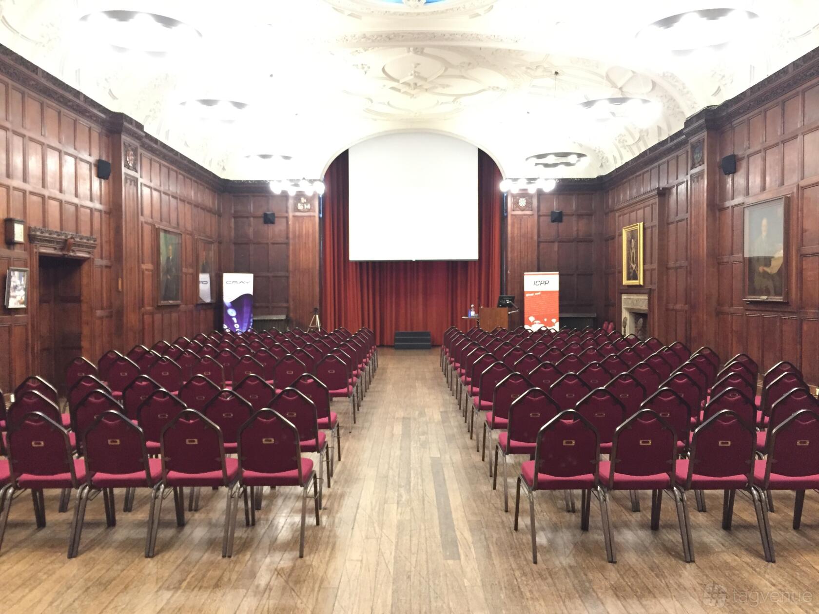 An event space with wood-paneled walls, ornate ceiling, and rows of red chairs facing a projector screen at Wills Memorial Building.