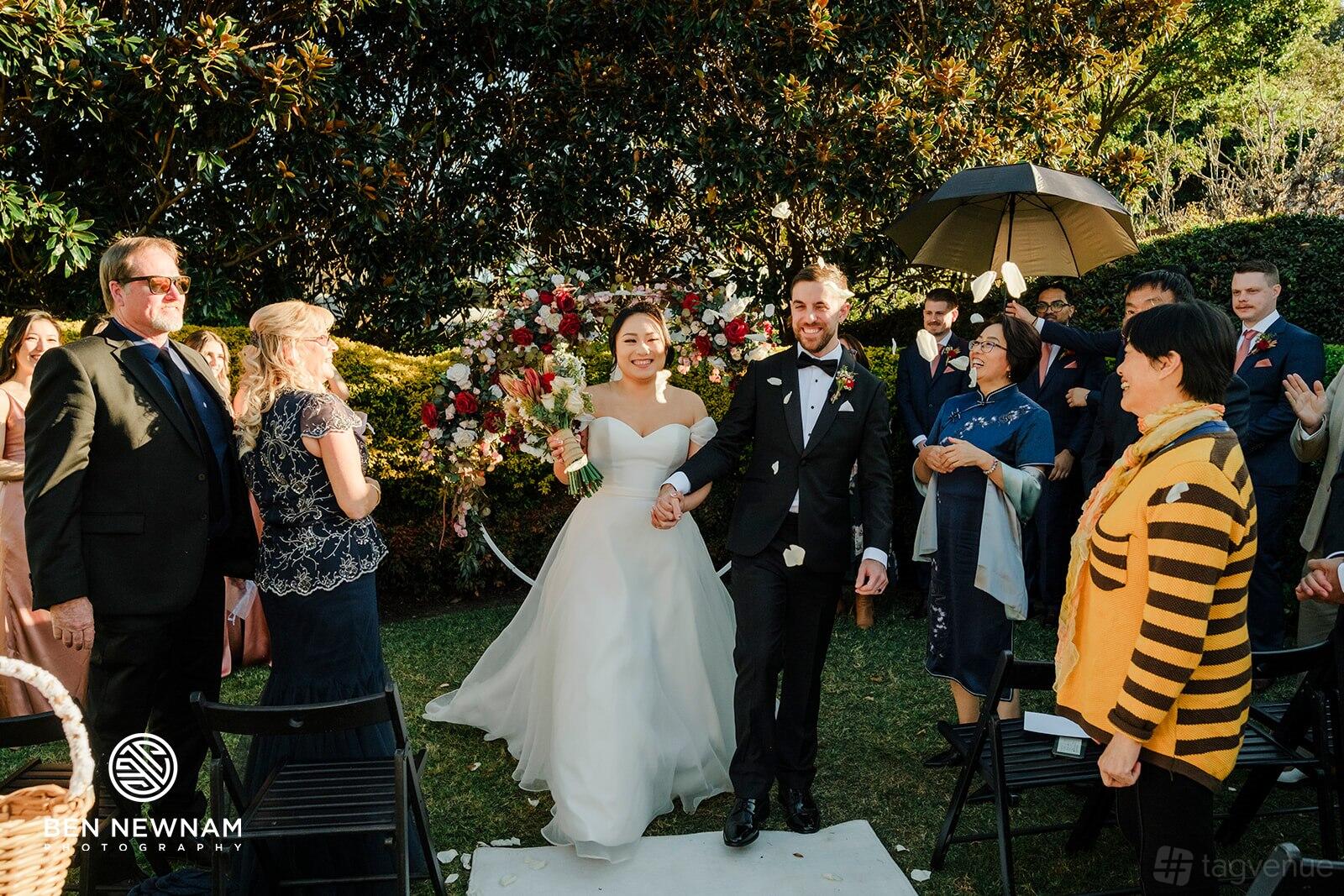 An outdoor event venue with a grassy aisle, floral arch, and guests seated under large trees at Will & Mike's.