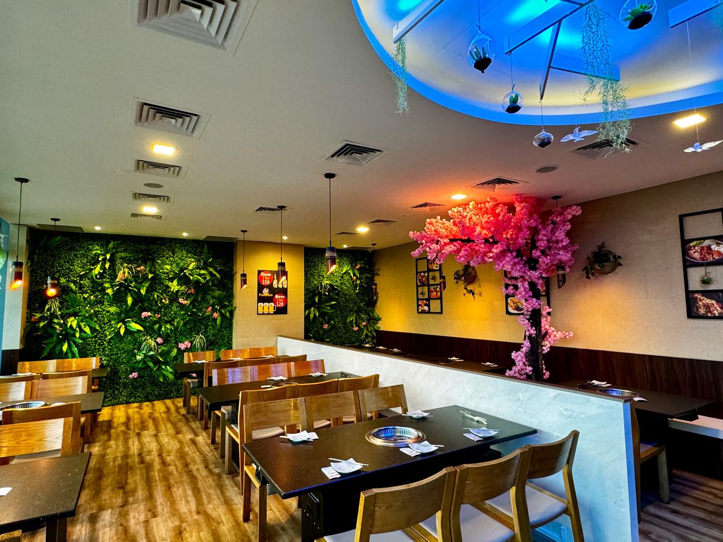 A restaurant with wooden tables, a pink cherry blossom tree, and lush green wall decor at Tenkaichi Japanese Restaurant.