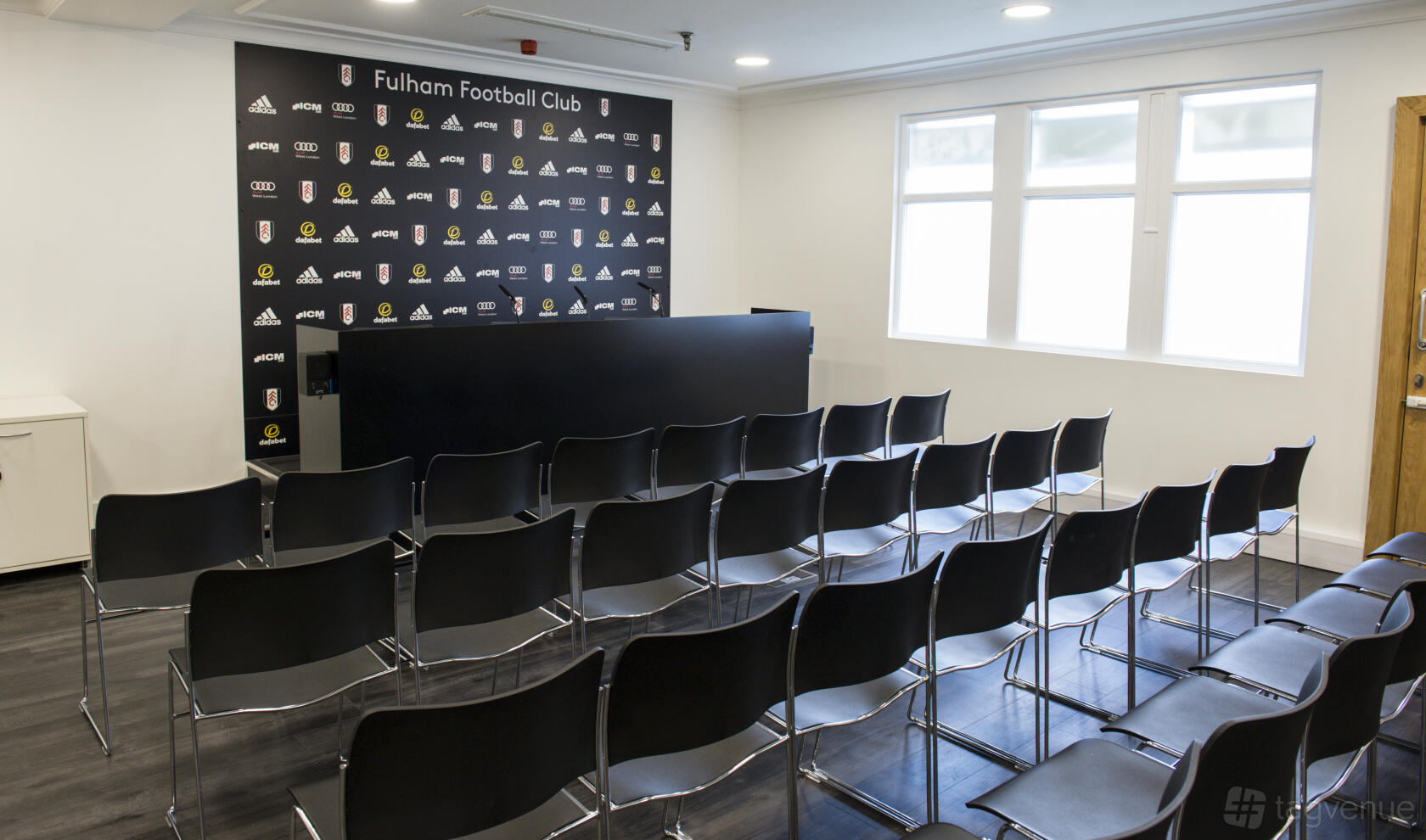 A stadium press room with a backdrop wall, rows of black chairs, and large windows at Fulham Football Club.