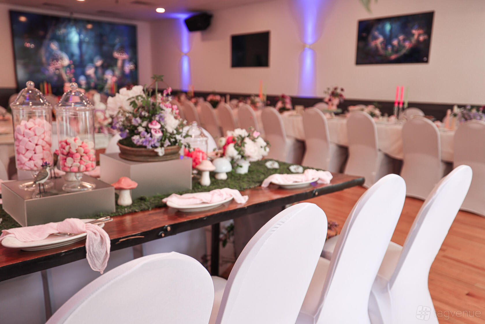 A hall with banquet tables covered in white linens, floral centerpieces, and pink candy jars at The Space Next Door.